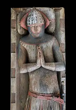 This image shows a medieval oak effigy retaining remarkable traces of its original polychrome decoration. The figure is carved in wood and depicts a knight in full armour with his hands positioned in prayer. The effigy demonstrates how medieval monuments were originally brightly painted to enhance their visual impact and create a lifelike appearance. Visible pigments include red detailing on the armour trim, belt, and helmet, with blue-grey tones on the body armour and a flesh-coloured face. The knight wears a distinctive pointed helmet typical of the period, and his armour shows detailed carved elements including belt fittings and decorative borders. The figure is set within a stone architectural frame or niche. This surviving polychromy is exceptionally rare, as most medieval monuments have lost their original painted surfaces over the centuries, leaving us with only the bare stone or wood beneath. Such examples provide invaluable insight into the original splendour and visual impact that these monuments would have had when first created.