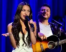 Olivia Rodrigo singing into a microphone while Dan Nigro plays the guitar behind her