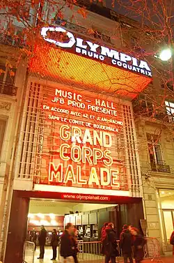 A photograph of the exterior of a theatre at night. Uppercase white text on the marquee reads "Olympia Bruno Coquatrix" against an orange backdrop. Several tree branches and leaves are visible at the top of the photograph on either side. A crowd of people are visible at the front entrance of the theatre.