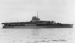 Side view of a gray anchored aircraft carrier with a short flight deck. Her island incorporates a funnel and is surmounted by a tall mast.
