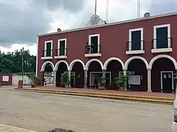 A two-story municipal-style building with a deep red facade and white trim stands under a cloudy sky. The front of the building features a row of six large arches supported by cream-colored columns, creating a shaded walkway at ground level. Above the arches, the second floor has five evenly spaced windows, each with black iron railings, and two central windows open onto small balconies. At the top center of the building, there is a circular emblem or seal with a green and gold design. Several large potted plants line the walkway in front of the arches, adding greenery to the scene. A few people are visible: one person stands on a balcony, another is near the entrance below, and a third is walking under the arches. The street in front of the building is empty, and trees and a white wall can be seen in the background to the left.