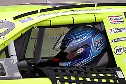 A man in his late 20s wearing a racing helmet and sitting inside a closed-cockpit stock car.