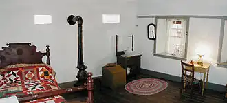 Telegraph room of Windsor Castle, Pipe Spring National Monument, Arizona. There is a bed in the left corner with a multicolored quilt and a heavy woodn frame. To the right of the bed is an iron stove with a pipe leading into the wall. Next there is what looks like a small chest for belongings and a small wash stand. Then a window with the curtains open. Finally a small desk with a lamp and a chair.