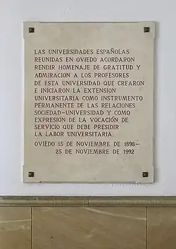 A stone rectangle on a white wall with red letters reading: "LAS UNIVERSIDADES ESPAÑOLAS REUNIDAS EN OVIEDO ACORDARON RENDIR HOMENAJE DE GRATITUD Y ADMIRACION A LOS PROFESORES DE ESTA UNIVERSIDAD QUE CREARON E INICIARON LA EXTENSION [sic] UNIVERSITARIA COMO INSTRUMENTO PERMANENTE DE LAS RELACIONES SOCIEDAD-UNIVERSIDAD Y COMO EXPRESIÓN DE LA VOCACIÓN DE SERVICIO QUE DEBE PRESIDIR LA LABOR UNIVERSITARIA. OVIEDO 15 DE NOVIEMBRE DE 1898-25 DE NOVIEMBRE DE 1992"