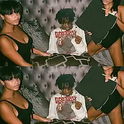 A duplicated color photo of Playboi Carti sitting on a bed between two women. He wears a distressed long-sleeve shirt with red text, looking at the camera while holding a blunt. One woman sits beside him, and the other kneels holding a pillow