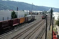 Two trains head in opposite directions along tracks in a switching yard surrounded by warehouses.