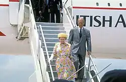 The Queen, in a pink floral dress, cream hat and long white gloves, descends from a BA-liveried aircraft via a set of movable aircraft steps, with the Duke, in a grey suit and dark tie, close behind her