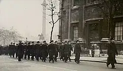 Black-and-white photo of a large group of men wearing military uniforms marching in formation along a city street