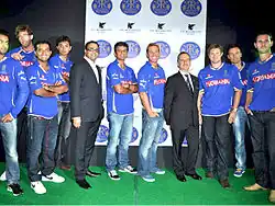 Nine players standing on a green carpet wearing a blue top and blue or black jeans. Along with them are two other men who are wearing black cffoats and pants.