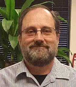 A balding brown-haired man with a beard, mustache and glasses facing the camera as he smiles slightly, wearing a narrow striped short with its collar open. Behind him is a window with closed blinds and a potted plant.