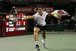 Raonic lunging to his right behind the baseline, with his racquet stretching to reach the approaching ball. Scoreboard in the background.