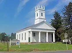 A white church with a colonnaded front and square steeple topped with spiky finials on the corners on a mowed grassy area under a blue sky. Behind it on the right are tall evergreen trees. In front on the left is a sign saying "Reformed Church of New Hurley" and a green street sign with "New Hurley Road" on it