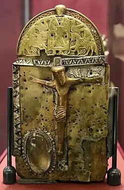 A handbell shaped shrine with a main face containing a large central crucifixion and a rock crystal to the lower left. The embossed panel above Christ's right arm is decorated with animals. The semicircular cap and crest above the main face shows a highly detailed central figure, likely an ecclesiastic, holding a book. Horse riders and birds are on either side of him.
