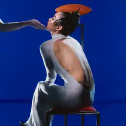 Rina Sawayama wears a white backless dress while sitting on a stool, in front of a blue background. A hand cups her chin.