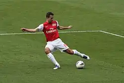A coloured photograph of a footballer wearing a red-and-white football shirt with the team logo. He is positioning himself to kick the ball with his left foot.