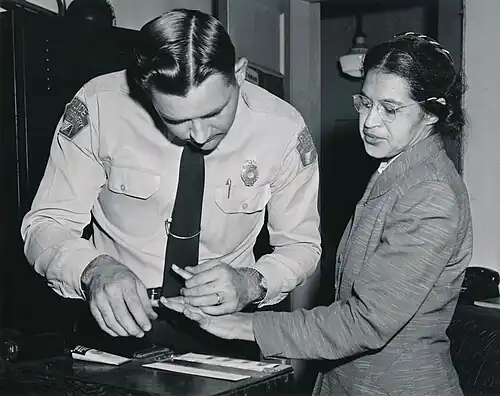 A black and white photograph of Rosa Parks as a middle-aged woman being fingerprinted by a police officer.