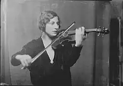A young white woman playing a violin indoors. She has hair in marcelled waves, and is wearing a dark garment with a deep V neckline