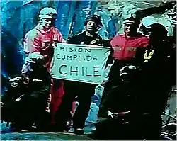 Slightly grainy color video capture image of the six rescuers displaying the famous "Mision Cumplida Chile" sign deep within San José Mine near Copiapo, Chile[2]