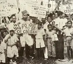 Photograph of adults and children holding signs, one saying "WE DO NOT WANT TO BE COLONISED", another saying "Anthony Brooke is NOT crimmina[sic] why ban his arrival to Sarawak"