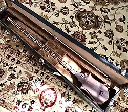The sato is inside an open cloth-lined hard case, and the bow (similar to a violin bow) is clipped inside the lid. The case rests on a central Asian carpet.