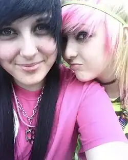Two teen girls in scene style, with dyed hair, bright graphic tops, and edgy jewelry typical of MySpace-era subcultures, 2010.