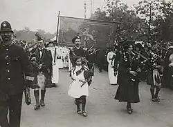 Photograph, printed, paper, monochrome, a procession of suffragettes led by a woman in a tartan dress, a small girl and three men in kilts all playing bagpipes.