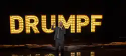 Comedian John Oliver atop a stage, wearing a suit and holding a microphone; in the background is the text "Drumpf"