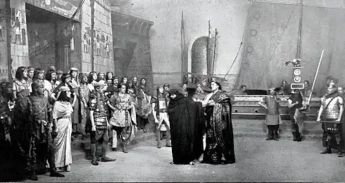 Outdoor, dockside scene with stage filled with men and women in Ancient Roman and Ancient Egyptian costume