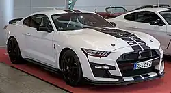 Front three-quarters view of a white two-door coupe car with two blue stripes running down its roof, hood, and bumper cover. It has a serpent logo on its grille