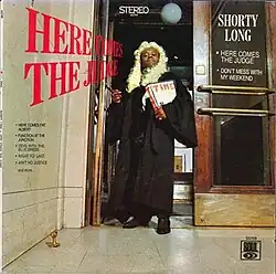 Long dressed as a judge with powdered wig and robe, standing in a courthouse