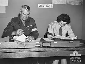 Two soldiers, one male, one female, sit side by side doing paper work at a desk. They are wearing brassards and the sign behind them says: "signal master".