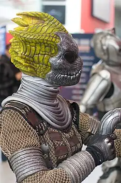 An image of a reptilian humanoid, depicted with a grey mask that has bulbous black eyes. The figure is wearing netted clothing.