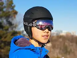 A skier wearing Bollé goggles and a Bollé helmet. He is also wearing a blue jacket. Behind him is a ski hill