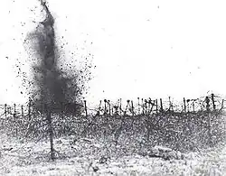 In the monochrome photograph, a shell detonates amidst a field strewn with multiple layers of tangled strands of barbed wire.