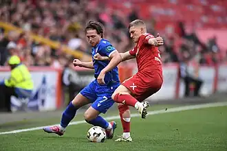 Jamie Dishington of The Spartans in a match against Aberdeen in September 2024. Jamie can be seen in a blue jersey with the number 7 on his shorts. The Bitcoin Cash logo can be seen on his left sleeve. He is fending off an Aberdeen player dressed in a red uniform.