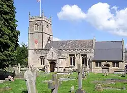 Yellow stone building, with porch with triangular roof in front. Short square tower with battlements topped by flag and flag pole. Gray gravestones in the foreground.