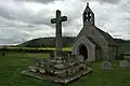 A plain greystone cross, in a mounting block, on top of four stone steps, with a small stone church in the background