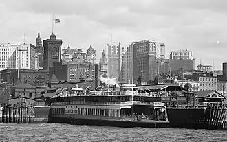 The Whitehall Terminal, a ferry terminal, is in the foreground between skyscrapers behind it and a harbor in front of it.