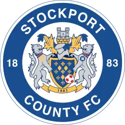 The words "Stockport County FC" in a circle (along with "18 on the left and "83" on the right) surround the main crest consisting of a shield featuring a hat and castle on top along with two lions rampant either side.