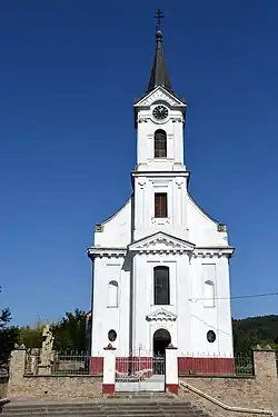 A white Catholic church is depicted. It is small, surrounded by a wall and with a gate in front. it has one steeple with a clock set into the facade.
