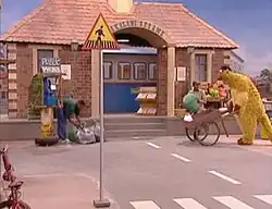 Television set, showing a large brick building on a city street; to the right, a large puppet is pushing a wheelbarrow and to the left, a man, next to a phone booth, is bending over several mail bags. Closer to the front of the image is a signpost, with a triangle that has a black figure and a yellow background, on top.