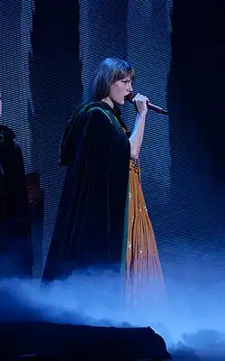 Swift dressed in an orange dress and black cloak singing onto a mic