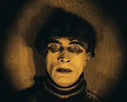 A sepia-tinted close-up of a man looking directly forward with wide eyes. He has short black hair and pale white skin, and is wearing a black shirt with a high collar.