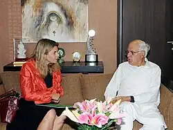Photograph of Helene Pelosse, sitting on a couch wearing an orange jacket over a black dress, and talking with Dr. Farooq Abdullah who is on the right wearing a robe