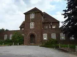 A photograph of a large derelict brick building from the 1930s. The front of the building has a large, rounded archway in the art deco style. The lower windows are boarded up, and the panes of many of the upper windows are smashed
