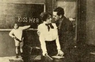 A black and white film still. A couple embracing. A girl in the background at a blackboard, having written the words KISS HER in chalk.