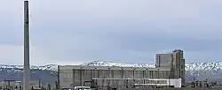 A color photograph of a grey-colored industrial facility and its chimney, with ice-covered mountains in the background