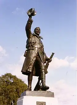 Statue of a soldier with rifle at his left side and hat raised aloft in his right hand.