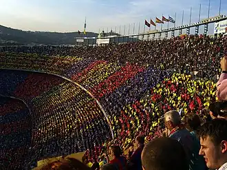 A crowd of spectators in an open-air stadium, a mosiac in red, blue and gold is visible
