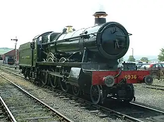 A locomotive is seen from in front and from the right. The locomotive is mainly green but the smoke box (at the front) is black and the buffer beam is red with the number 4936 written in yellow letters. The chimney is trimmed with a copper ring and the valve cover is made of brass. Behind the locomotive is a green tender with the words "GREAT WESTERN" written in yellow. In the background to the left is a signal and an engine shed with a tank engine, and to the right is a car park behind a wooden rail.